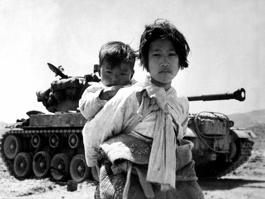 A Korean refugee girl trudges past a stranded M46 tank in Haengju, Korea, carrying her younger brother on her back